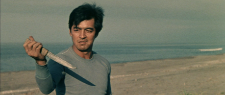 A Japanese man in his 30s wearing a casual shirt and standing in front of a lake holds a dagger in his hand, and is smiling at it.