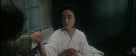 A Japanese woman sitting up in bed, wearing a white bathrobe and holding an apple, takes the hand of a man standing beside the bed (back to camera) 