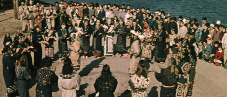 A shot from above of a large group of indigenous Ainu, wearing colorful costumes, performing a ceremonial dance in the open air