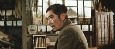 A middle-age Japanese man sits in the foreground facing left and looks at someone out of the frame with a baleful stare