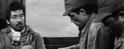 A bearded man, sitting in a train and wearing an army uniform, warily watches his two seat mates, who are also wearing uniforms but who are both looking down and away from him