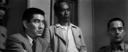 Three Japanese men, two sitting and one standing in the middle, stare unsmiling at someone or something unseen