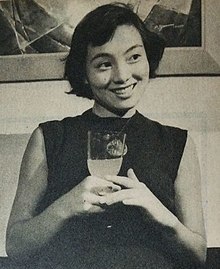 A young Japanese woman in modern (1950s) clothing, holding a drink in her hands and smiling at something off-camera