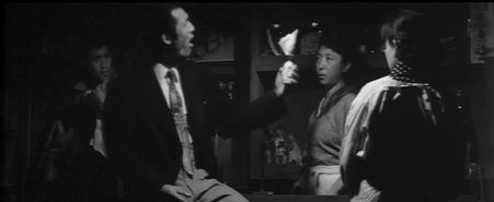 A tall young Japanese man wearing a suit holds aloft a small jar with cash stuffed into it, as a worried-looking middle-aged woman and Yae (back to camera) watch