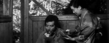 A woman on a local train, standing, watches a man in rages eating a rice ball