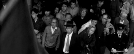 Among a Japanese crowd, a man wearing a suit and hat, Yumisaka, stops to watch a street protest, of which one of the flags can be seen