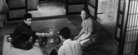 A Japanese family, a mother and two sons, sit on tatami mats around a table, as the older boy hands the young one money, to the delight of the mother