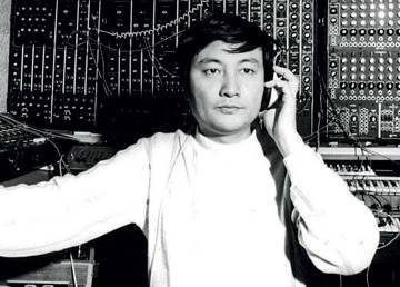 A young Japanese man in modern clothes, surrounded by electronic equipment and wearing headphones, gazes distractedly in front of him 