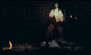A young man in ancient Japanese clothing lies sleeping on the ground, while a young woman in white stands over him; there is a small fire on the ground to the left of the man