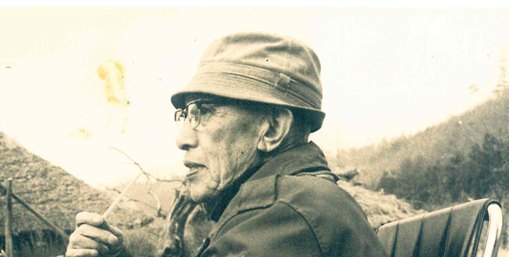 An old Japanese man wearing a cap, sitting outdoors on a chair and holding a cigarette in his hand