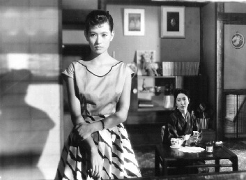 In a modern (1950s) Japanese house, an attractive young woman in modern dress, standing, looks towards the camera, while behind her and to her left, seated at a table and wearing kimono, a slightly older woman looks at her 