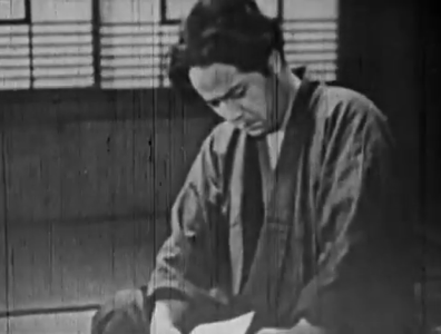 In medium shot, a young Japanese man in traditional dress sits in a room, looking downcast