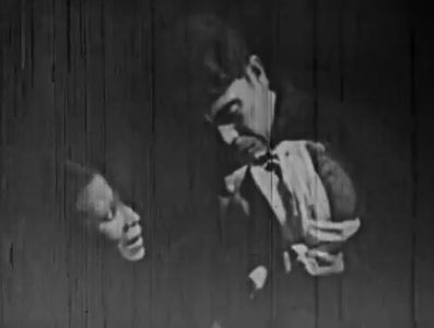 In near-total darkness, an elderly woman and a young man look at the corpse of a man with a cloth around his bleeding head