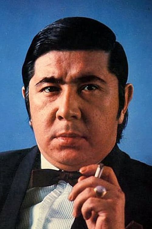 Color photo of a Japanese man in his late 30s, wearing a suit and a bow tie and with a cigarette in his hand