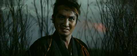 In a field, a young samurai with a headband looks into the camera with an intense expression