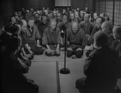 A group of poor Japanese farmers from the feudal era are gathered kneeling or sitting in a room, with the focus of the men upon one old man in the right corner of the frame