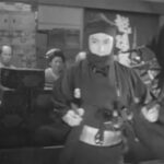 A masked man in ninja costume, carrying swords, points pistols in the direction of the viewer; he is surrounded by similarly-dressed henchmen, while several Japanese in feudal period clothes watch them nervously