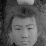 A Japanese boy from the feudal era, standing with his back to a tree, starting directly towards the camera, with a piece of fruit on top of his head
