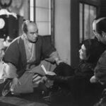 Near a window, a middle-aged Japanese man in samurai costume, in a kneeling position, looks with concern at a man in a man lying in the floor with his hand outstretched to him, while two other men also watch with concern