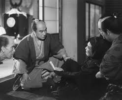 Near a window, a middle-aged Japanese man in samurai costume, in a kneeling position, looks with concern at a man in a man lying in the floor with his hand outstretched to him, while two other men also watch with concern