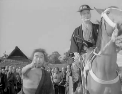 The same man with the rifle from the previous picture sits on horseback on the right of the frame, while the man who had bowed stands on the ground on the left, pointing at something in the distance, as a group of men watch in the background