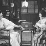 A young Japanese couple from the early 20th Century, shown relaxing at home, she playing a wind-up gramophone; he wearing a bathrobe and smoking a cigarette