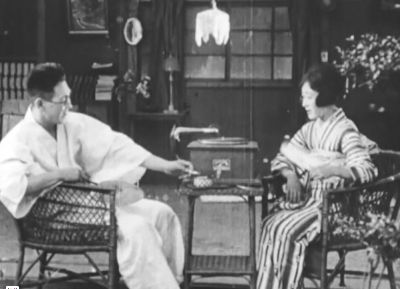 A young Japanese couple from the early 20th Century, shown relaxing at home, she playing a wind-up gramophone; he wearing a bathrobe and smoking a cigarette