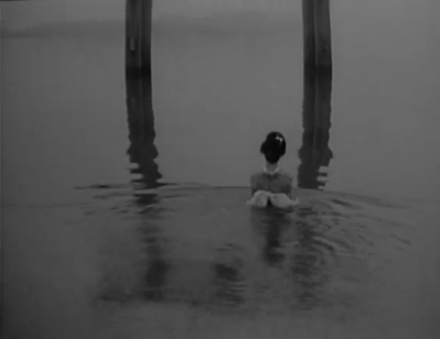 A photo of a woman in traditional Japanese dress standing in the middle of a lake, almost up to her shoulders in the water, and flanked by two large pillars which cast their reflections in the water