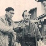 At left, in the middle distance, an elderly Japanese man wearing modern (1950s) clothing, including a beret, stands in the street, addressing a man in feudal period costume standing next to him