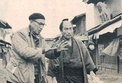 At left, in the middle distance, an elderly Japanese man wearing modern (1950s) clothing, including a beret, stands in the street, addressing a man in feudal period costume standing next to him
